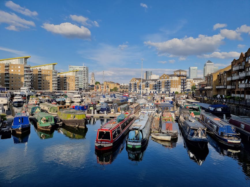 A picturesque courtyard garden at Limehouse Basin, featuring a narrow waterway with floating buoys and water lily pads, bordered by a well-maintained grassy edge. Behind the water, there is a paved ga